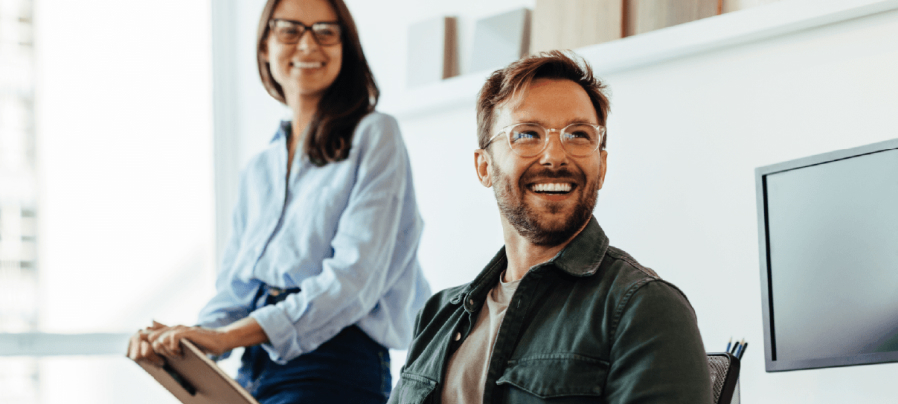 Man with glasses laughing at something out of view with a woman in glasses sits behind him smiling.
